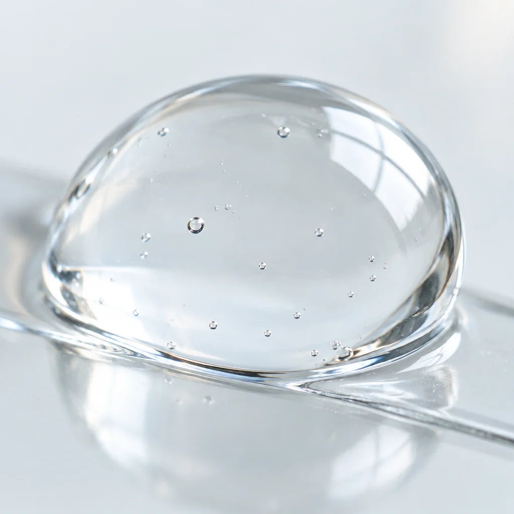 An extreme macro photograph of a water-clear skincare serum bead resting on a very thin transparent